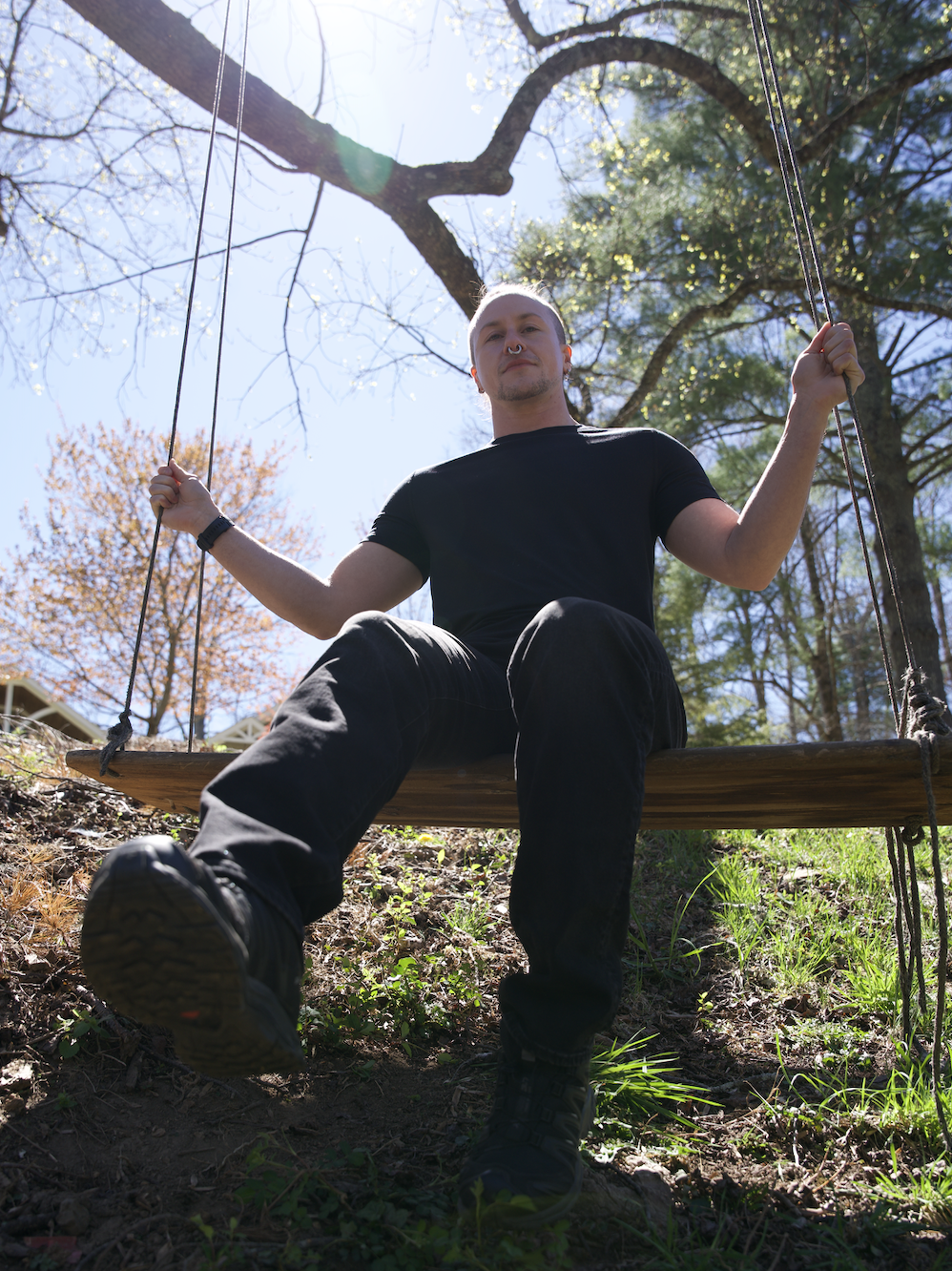 headshot of Sirus, a white person in black pants and shirt, sitting on a swing attached to a treebranch in the sunlight